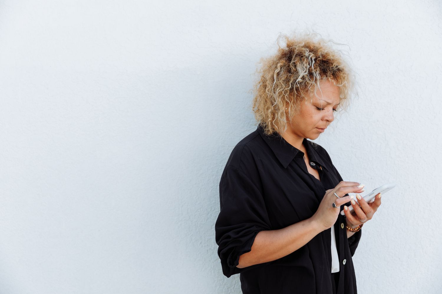 A woman with curly blonde hair leans against a pale wall, reading her phone with a tired, focused face