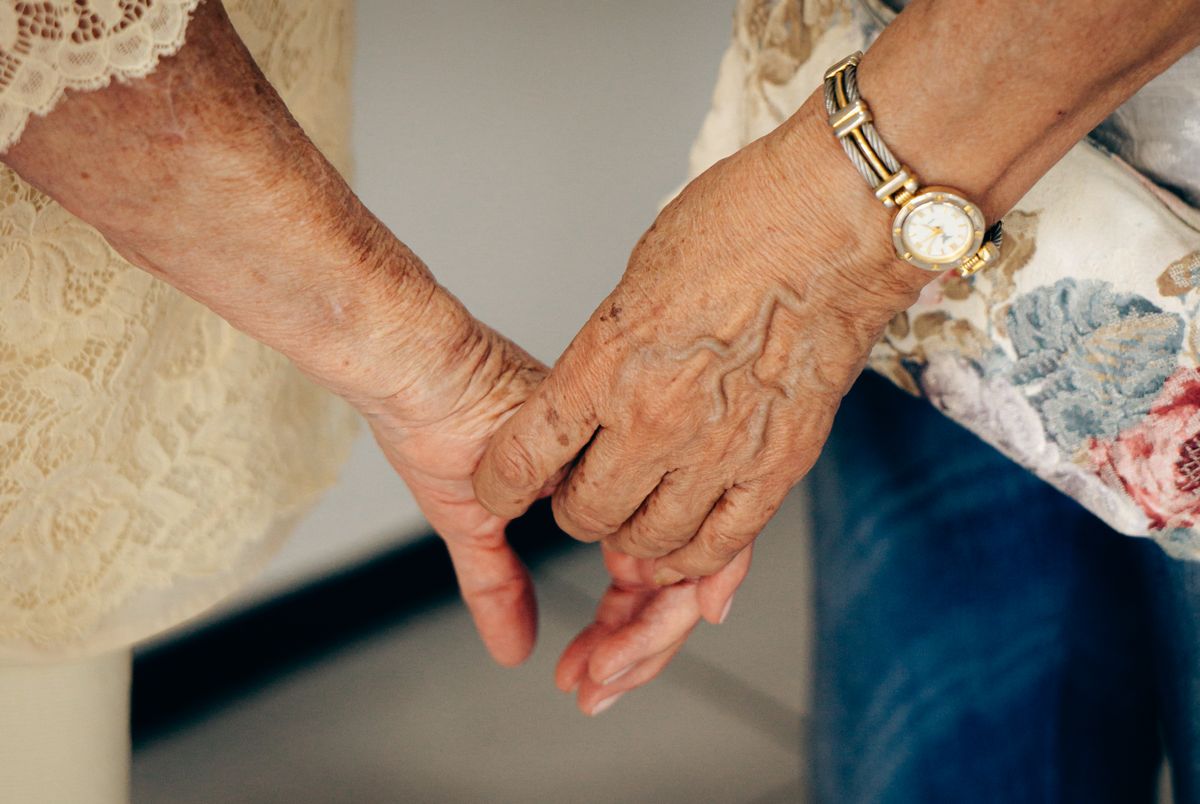 Two older hands clasped together, one wearing a gold wristwatch
